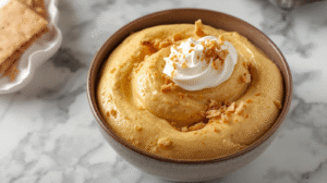 A bowl of fluffy pumpkin mousse topped with whipped cream and graham cracker crumbs, surrounded by autumn decorations.