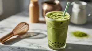 A tall glass of matcha milk tea with layers of green foam sits in bright sunlight, surrounded by blurred matcha powder and a whisk in the background. The creamy drink shines with fresh, earthy tones.